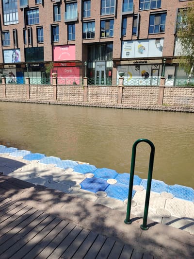 This image captures a vibrant urban waterfront scene in Eskişehir, Turkey, under bright daylight. In the foreground, a wooden boardwalk transitions into a floating modular dock made of interconnected blue, white, and light gray plastic hexagons. A green, U-shaped metal handrail is secured at the edge of the dock, partially submerged in the water. Shadows from overhead trees or structures fall across the wooden planks and the floating dock.
The midground features a calm, murky brown river or canal, stretching across the frame. On the opposite bank, a textured stone retaining wall supports a light green metal fence adorned with ornate lamp posts.
In the background, a multi-story brick building with numerous windows dominates the skyline. The ground floor of this building houses several commercial establishments. Visible signs include "QNB" with the text "QNB'ye geçiyorum." and an image of a smiling woman. Another prominent sign reads "Penti" with "ENJOY BEING YOU" on a pink background, and another red "Penti" sign below it. Further along, a branch of "Yapıkredi" bank is visible. On the right, "odeabank" is seen, featuring a logo with "odes" and text like "Geniş ürün portföyü ile yatırımın merkezinde sen varsın." and "Hedeflerine uygun yatırım seçenekleri ile..." The building itself is identified as "ALÇIN KILIÇOĞLU PLAZA". The clear skies and strong shadows indicate a sunny day. No people are actively visible, only images on advertisements.