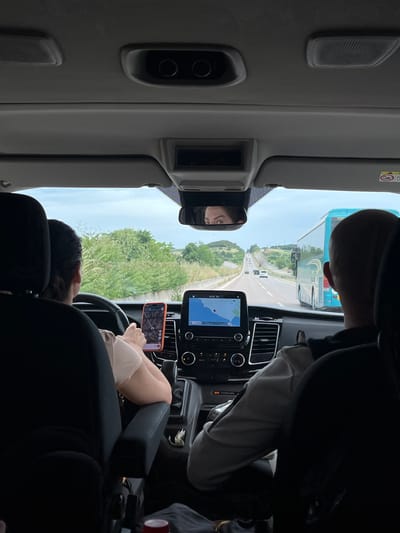 A photograph taken from within a car interior shows a partial view of two individuals during a trip. The driver holds an orange smartphone, with a map visible on the screen, positioned near the steering wheel. A screen on the vehicle dashboard displays a map as well. The rearview mirror reflects the driver's face. Through the windshield, a bus is visible. The location appears to be on a road near Άγιος Παύλος, Greece, as indicated by the road scenery.
