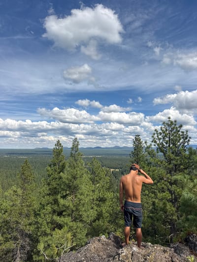 A male subject is depicted from behind, standing on a rocky outcrop overlooking a vast forest landscape, located in Three Rivers, United States. He is shirtless, wearing dark shorts with a light-colored stripe on the left side, and a backward-facing tan baseball cap. His left hand is positioned on the cap. The foreground consists of dark, irregular rock formations. Immediately beyond the subject, several tall, dark green coniferous trees rise. The mid-ground and background are dominated by an extensive, undulating evergreen forest canopy, extending towards a faint line of distant mountains or hills on the horizon. The sky above is bright blue with numerous scattered white cumulus clouds of various sizes. The scene is illuminated by daylight.