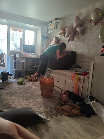 The image depicts a cluttered indoor scene. A woman in a green t-shirt and dark pants is leaning over two dark brown storage containers stacked on a white daybed. The daybed is covered with a white sheet and various items of clothing. To the left of the daybed, a grey cat tree with two levels is visible, with a white blanket draped over the top level. A black trash can is located to the left of the cat tree, with crumpled white paper scattered on the floor. A green plush frog toy lies on the patterned rug in front of the cat tree. A large orange paper bag is situated to the right of the frog toy. Further to the right, a small doll lies on its back near a brown backpack. The room features a window with blinds and a desk with a computer monitor. Balloons and decorative fabric bows are attached to the wall in the background. The floor is a mix of patterned rug and light-colored wood planks. The overall environment appears to be a bedroom or living area that is in disarray. There is no information in the image that allows for the determination of the location in the city of Berdsk municipality, Russia.