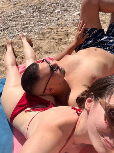 Captured from an overhead perspective, the scene unfolds on a sandy beach, likely situated in El Poble Nou de Benitatxell / Benitachell, Spain. A couple is the central focus, lying on the sand in a sunbathing position. The man is clad in dark blue swimwear with the brand name "Calvin Klein" imprinted in a repetitive pattern. He is lying on his back, with his head positioned towards the top right corner of the frame. Sunglasses are perched on his nose. The woman is positioned close to the man. She is wearing a red bikini. A light blue towel lies beneath them. The sand is comprised of both fine grains and small pebbles. The lighting suggests a bright, sunny day.