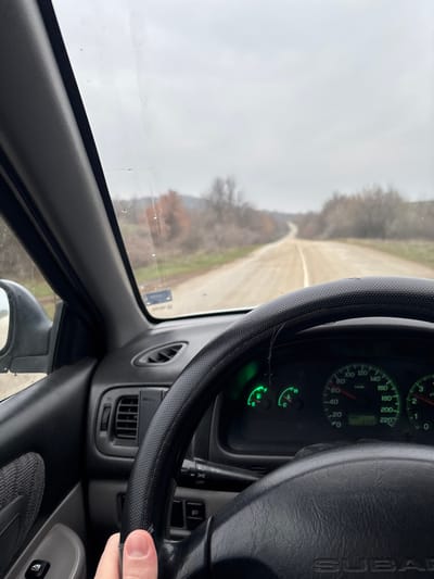 The scene is captured from the interior of a Subaru vehicle, looking forward through the windshield onto a rural road in Suhache, Bulgaria. The driver's right hand, with a trimmed fingernail, is positioned at the 7 o'clock position on a black, perforated steering wheel cover, obscuring the lower left portion of the steering wheel, where the "SUBARU" logo is partially visible.
The instrument cluster, visible above the steering wheel, is illuminated with green backlighting, displaying a speedometer, tachometer, fuel gauge, and temperature gauge. The odometer reads "27242". A control stalk, likely for lights, extends from the steering column. A black phone mount is attached to the dashboard, adjacent to an air vent on the left. The interior door panel, featuring a grey fabric insert and a window control switch, is visible on the far left, alongside the exterior rearview mirror, which reflects portions of the overcast sky and roadside. The A-pillar runs vertically along the left edge of the windshield.
The windshield itself appears dirty, with smudges, streaks, and minor surface imperfections. Beyond the windshield, a light brown, unpaved or gravel road with a faded central white line extends into the distance, curving gently. The road is bordered by green grass and sparse, deciduous vegetation, including trees with bare branches and some with reddish-brown leaves, suggesting a late autumn or early winter setting. The sky is uniformly overcast and light gray, indicating a cloudy day. The horizon in the distance reveals gently undulating terrain.
