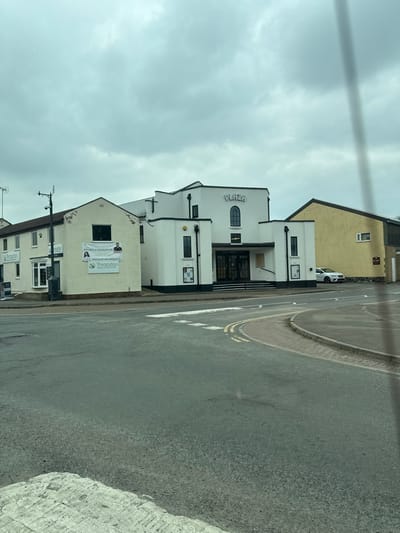 The image depicts a street scene in Thrapston, United Kingdom, featuring the Plaza building at 41 High Street, situated at a road junction. The central subject is the Plaza building, a white, two-story Art Deco-style structure with dark accents. The name "PLAZA" is prominently displayed in raised letters above a central arched window on the upper facade. The lower level features a recessed main entrance with dark double doors, accessible via several white steps. Flanking the main entrance are two symmetrical vertical sections, each with a narrow dark-framed window and poster display frames at ground level.
To the left of the Plaza building stands a lighter-colored, multi-story building identified as "Thrapston Dental Centre," displaying banners on its side that advertise "IMPLANTS & PERIODONTICS" and "SPECIALIST ORTHODONTICS." A black street lamp post with attached antennas is positioned in front of this building. To the right of the Plaza, a simpler, two-story yellow building with a brown roof is visible, behind which a white sedan is parked.
The foreground consists of an asphalt road surface with various road markings, including white lines for a crosswalk and double yellow lines along a curved curb, indicating a road intersection or turning area. The sky above is uniformly overcast with no direct sunlight visible. No individuals are present in the frame.