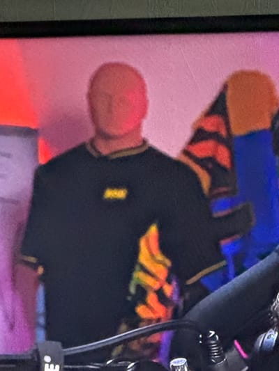 A close-up, slightly blurry, eye-level shot shows a person from the chest up, wearing a black t-shirt with yellow trim and a yellow logo on the left chest. The person is bald and has a reddish hue to their skin, possibly due to lighting. Behind the person, there is a colorful, abstract background with areas of pink, red, orange, yellow, and blue. To the right of the person, a large object with orange and black stripes and a blue section is visible. In the foreground, the edge of a black object, possibly a camera or microphone stand, is present, with some metallic and black details. The overall impression is of a person standing in front of a vibrant and possibly artificial backdrop. The image is undated and its specific location in Gothenburg, Sweden, cannot be determined from the visual information provided.