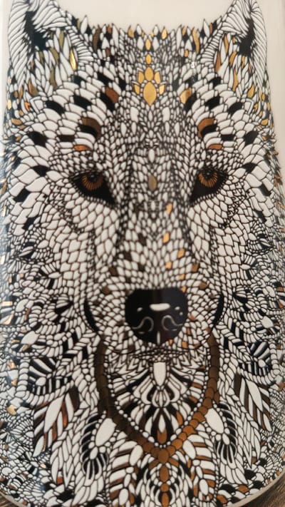 The image is a close-up, top-down view of a white ceramic object, likely a mug or vase, decorated with an intricate black, white, and gold pattern that forms the face of a wolf. The wolf's eyes are a warm brown, with a dark pupil. Its nose is black and heart-shaped, with a lighter colored detail on the bridge. A decorative collar-like band, composed of interconnected gold and black shapes, encircles the wolf's neck area. The fur is depicted with a repeating scale-like pattern, with some scales highlighted in gold and black. The lighting suggests an indoor setting, and the object appears to be placed on a flat surface, though the surface itself is not clearly visible. There is no indication of the city of Zvenigorod, Russia, or any other location.