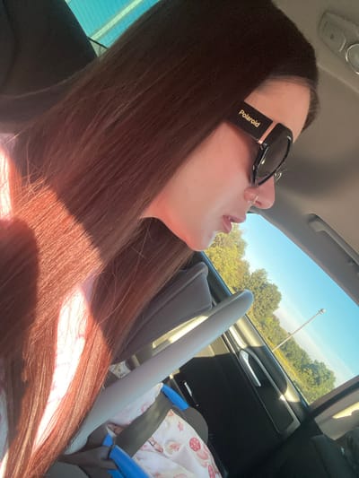 A woman with long, dark hair is sitting in the passenger seat of a car, looking out the window. She is wearing black sunglasses with "Polaroid" written on the side. Sunlight streams through the car's sunroof, illuminating her hair. In the background, green trees and a blue sky are visible. The interior of the car, including a car seat and a portion of the steering wheel, is also visible. The scene appears to be set in a rural or suburban area. It is not possible to determine the exact location of Herasymivka, Ukraine from the image alone.