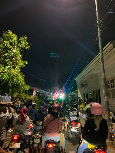 The image depicts a crowded street scene at night in the city of Kedungbanteng, Indonesia. The perspective is from behind several individuals on motorcycles, facing forward into a dense gathering of people and activity.

In the immediate foreground, the backs of four individuals on three motorcycles are visible. All three motorcycles have illuminated red taillights. The leftmost rider wears a red headscarf, a light-colored top, and dark pants, on a motorcycle with a license plate partially visible as "T 47 SG." The middle rider wears a pink top and light-colored jeans, on a motorcycle with a license plate "S 2110 O." The rightmost rider wears a pink helmet, a dark jacket, and a light-colored head covering, on a motorcycle without a visible license plate. To the far left, a person carries a wooden frame laden with multiple plastic water bottles on their left shoulder.

The narrow street ahead is filled with a large crowd of people, a mix of pedestrians and individuals on various motorcycles, all facing towards the background. An Indonesian flag (red and white) is visible on the left side of the street amidst the crowd. On the left side of the street, a large tree with green foliage is prominent.

Further down the street, bright artificial lighting, predominantly green and white, illuminates an apparent event or stage, with light beams projecting into the night sky. Blue and purple string lights are strung across the street overhead. On the right side of the street, residential buildings with white facades are visible, one featuring an external air conditioning unit. Numerous overhead utility lines crisscross the upper portion of the frame. In the dark night sky, above the street, a distinct pattern of green laser lights is projected.