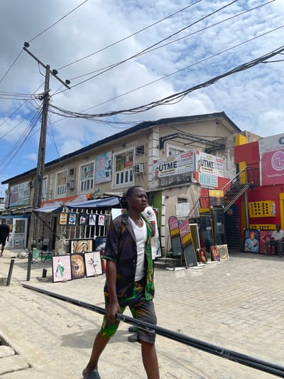 This is a street scene in Lagos, Nigeria, likely in a commercial area. A man walks towards the right of the frame, carrying a long, dark, cylindrical object horizontally. He is wearing a dark patterned shirt over a white undershirt and patterned shorts. In the background, a multi-story building with various signs and advertisements is visible. Some signs advertise educational services, such as "UTME" and "IELTS". There are also displays of framed pictures and mirrors on the sidewalk in front of the building. Power lines and a utility pole are prominent in the sky above. The ground is paved with interlocking bricks. To the far right, a man is seated on a chair outside the building. The sky is partly cloudy.