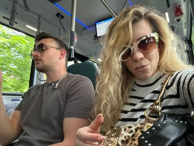 Two people are seated on a bus. A man on the left wears sunglasses and a grey t-shirt. A woman on the right wears sunglasses, a striped shirt, and has a patterned bag. The bus has a blue light strip and a display showing the route, potentially the 147 station, which includes Dronten, Netherlands. A security camera is visible on the bus ceiling. The view out the window suggests the bus is in motion.