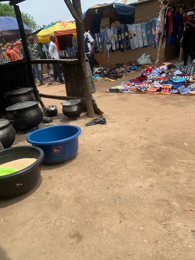 This media file captures an outdoor market scene in Nigeria, likely during daytime with clear weather conditions. The foreground is dominated by a collection of large cooking pots of various sizes, some with lids, arranged on the sandy ground. To the right of the pots, a large blue plastic basin stands prominently. Next to it, a black plastic basin is filled with what appears to be uncooked rice, with a bright green rim.
In the background, a stall is set up with a variety of clothing items displayed. Some are neatly hung on a wooden structure, while others are piled up. The clothing appears to be a mix of casual wear, including jeans and patterned tops. Several people are visible within the market. One person, wearing a white shirt, stands near the stall, while another, in an orange patterned shirt, is seated further down. A person in a blue and white striped shirt is also visible in the upper left quadrant.
A yellow umbrella with red and white branding is visible, with text that reads "MAGGI" in large letters, repeated. Below that, smaller text is visible, including "Cook The Difference". The overall impression is a bustling local market with goods for sale, including food items and clothing, and evidence of daily life activities.