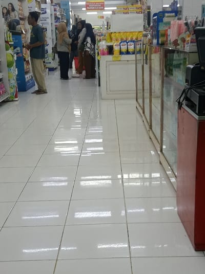 An interior scene depicts a supermarket or large retail store located in Nganjuk, Indonesia. The floor consists of glossy white ceramic tiles, reflecting rectangular overhead lighting fixtures. In the mid-ground, five human subjects are present. On the far left, one male subject wears a dark short-sleeved shirt and light-colored long pants, standing adjacent to a tall product display unit that features a promotional graphic. To his right, four female subjects are positioned near a white display counter. Three of these females wear headscarves; one in a light-colored headscarf and light top, another in a dark headscarf and light top, and a third in a dark headscarf and patterned top with dark pants and light footwear. The fourth female subject, partially obscured, also wears a headscarf. The white counter holds numerous yellow-packaged items. Above the counter, a red banner displays white Indonesian text. On the right side of the frame, a multi-paneled glass display cabinet with light-colored framing extends towards the background. The extreme right foreground shows a segment of a red service counter, topped with dark cables and electronic equipment. The background contains additional retail shelving and commercial signage. The subjects are generally stationary.