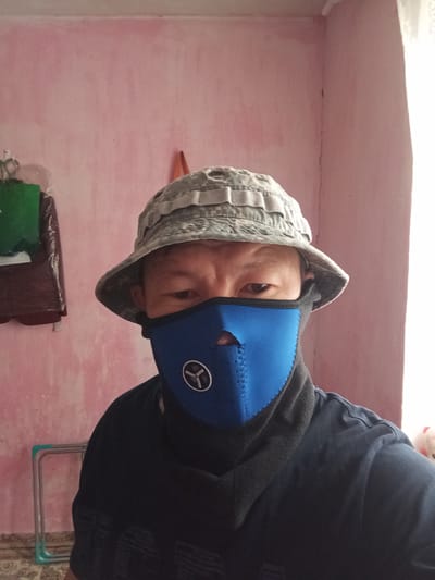 A person is pictured wearing a camouflage-patterned bucket hat and a blue face mask, with their head tilted to the side. They are also wearing a dark blue hooded jacket. The background appears to be an interior space with a pinkish wall. To the left of the person, a portion of a metal and green plastic shelf or rack is visible. In the upper part of the image, there are some stacked items, including a dark brown bag and a green object, possibly a bag or container. The lighting suggests it is daytime, and no specific weather conditions are indicated. The overall impression is casual and informal. No visible text is present in the image.