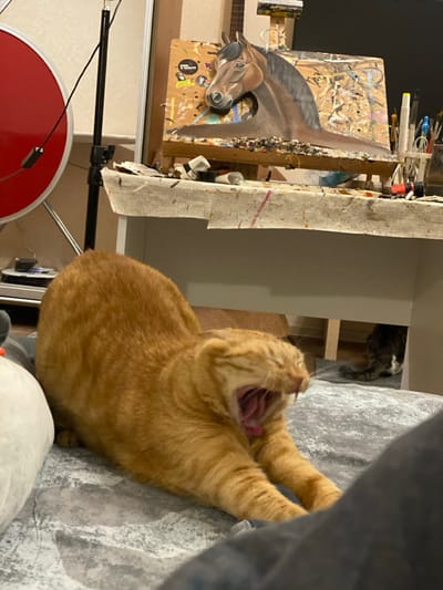 This image captures an indoor scene, likely an art studio or creative workspace, located in Inkerman, Ukraine. In the foreground, an orange tabby cat is stretching forward, extending its front paws and yawning widely, displaying its open mouth with visible teeth and tongue. To the immediate left of the orange cat is a partially visible white, soft object, possibly a cushion or plush toy. The cat is positioned on a grey fabric surface with a subtle pattern. On the far right, a dark-colored trouser leg is partially visible.
In the mid-ground, a white table is present, covered with a paint-splattered, light-colored cloth. An easel stands on the table, holding an acrylic or oil painting of a brown horse's head and part of its neck, depicted against a multi-colored, abstract background. The easel itself shows signs of extensive paint splatters. Various art supplies, including paint tubes and brushes, are scattered on the table surface next to the easel.
In the background, on the far left, a red, circular studio reflector or light accessory is visible on a stand, next to a black vertical pole. Below the white table, a second, dark tabby cat is partially visible, appearing to be resting on the floor. The overall lighting suggests an interior setting, possibly illuminated by artificial light.