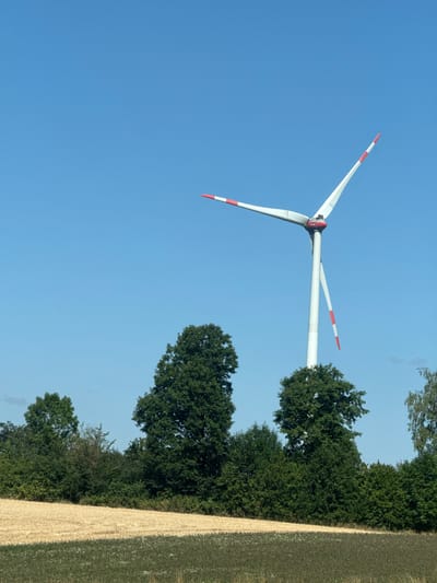 The image depicts a wind turbine against a clear blue sky, located in Liederbach, Germany. The wind turbine, predominantly white with red accents on its blades, stands tall with its blades extended. In the foreground, a field of dry, golden wheat is visible, bordered by a strip of green vegetation. Behind the wheat field, a dense line of lush green trees forms a natural backdrop, partially obscuring the lower section of the wind turbine. The overall scene is rural and serene, capturing a typical landscape of the German countryside.