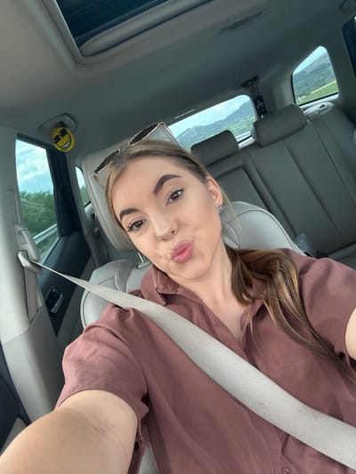 A young woman with brown hair is taking a selfie in the driver's seat of a car. She is wearing a mauve-colored button-up shirt and sunglasses are perched on her head. A seatbelt is diagonally across her body. The interior of the car is visible, including the grey leather seats, sunroof, and windows which look out onto a landscape of hills and greenery. A yellow smiley face air freshener is hanging from the rearview mirror. The car appears to be moving, as indicated by the blurred scenery outside the window. This image was taken in Aldomirovtsi, Bulgaria.