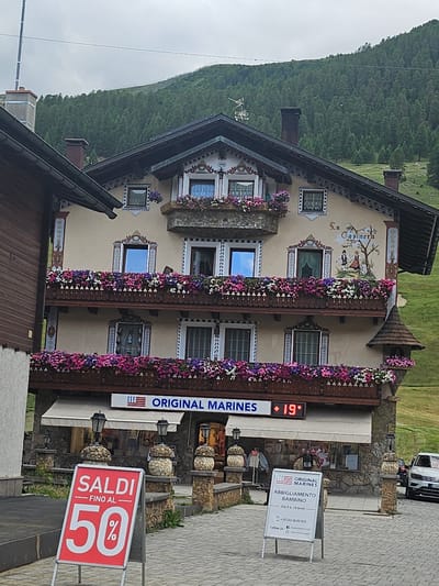 The image shows a multi-story building with ornate architectural details and balconies filled with flowers, located in Livigno, Italy. The building has a light-colored facade, dark brown trim, and a dark brown roof. Several windows with decorative frames are visible, and a mural depicting two figures and a tree is painted on the upper level. A sign above an awning reads "ORIGINAL MARINES" with a temperature display showing "19°". In front of the building, there are stone pillars with lamps and a red A-frame sign displaying "SALDI - FINO AL 50%". Another white A-frame sign to the right has text in Italian and an American flag emblem. A white SUV is parked on the right side of the image, and a wooded hillside is visible in the background under a cloudy sky. The scene appears to be a street or pedestrian area with a shop.