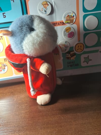 A stuffed animal hamster wearing a red hoodie with a white drawstring is the central subject of the image. It is positioned on a wooden surface, likely a table or desk, in a dimly lit room. Behind the hamster, a colorful chart or board is visible, featuring various circular stickers with images and text in Russian. One sticker depicts a person practicing martial arts, another shows a box of toys, and there's a sticker with a sad emoji. The chart also has numbers and text that appear to be part of a reward system or activity tracker. The overall scene suggests a child's play area or a learning environment. The image does not provide any information that can be used to determine its location in Pervomaisk, Ukraine.