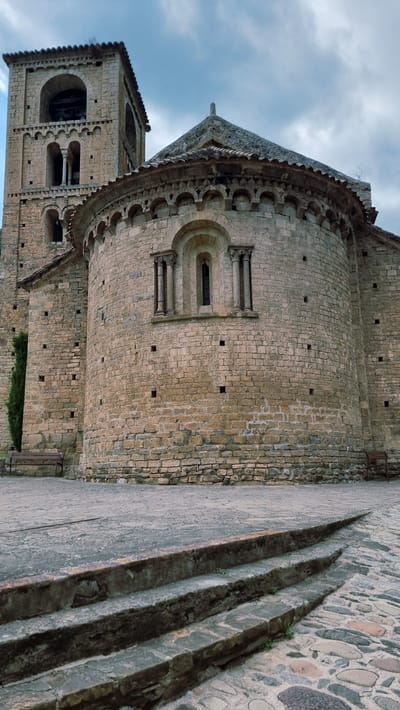 The image shows the exterior of a Romanesque church in Camprodon, Spain. The church features a tall bell tower with arched openings and a rounded apse adorned with decorative arches. The walls are constructed of weathered stone blocks. In front of the church, there is a paved courtyard with stone steps leading up to it. A narrow, dark green evergreen tree stands to the left of the apse, and a wooden bench is visible next to it. Another bench is partially visible to the right. The sky above is cloudy.