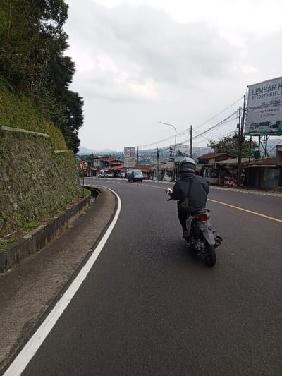 The image depicts a street scene in Ciloto, Indonesia, under an overcast sky. A dark-colored scooter, ridden by an individual wearing a dark jacket, dark pants, and a full-face helmet, travels away from the viewer in the right lane of an asphalt road. The road surface appears dark and possibly damp. A solid white line delineates the left edge of the road, bordering a concrete drainage ditch and a steep, vegetated retaining wall. Beyond the wall, dense green foliage is visible. Ahead of the scooter, the road curves slightly right, revealing additional vehicles, including dark-colored vans, in motion. On the right side of the road, numerous utility poles with overhead wires are present. A large billboard advertises "LEMBAH HIJA RESORT HOTEL & RESTAURANT." Below the billboard and further along the road, small buildings with corrugated metal roofs line the roadside. In the mid-ground, a dense cluster of buildings indicates a town or settlement, with a prominent tall, slender structure rising above the rooftops. Distant hills or mountains form the backdrop. A circular red prohibitory traffic sign is visible on the left side of the road in the mid-distance.