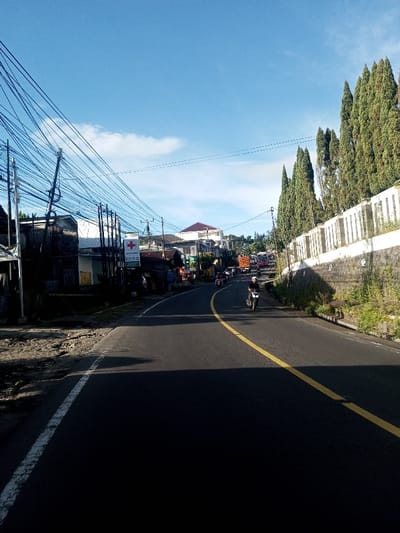 The image depicts a street scene in Khouathineung, Laos, under a clear blue sky with scattered white clouds. The paved road, marked by a solid white line on its left edge and a solid yellow centerline, curves gently to the right and appears to ascend slightly in the distance. Strong sunlight from the left casts long shadows across the foreground and mid-ground of the road.
On the left side of the road, a dirt and gravel shoulder transitions to a congested array of utility poles supporting numerous black overhead wires. Behind these poles are various low-rise buildings of differing construction, including one with a white sign displaying a red cross symbol, indicative of a medical facility or pharmacy.
The right side of the road features a segmented, light-colored concrete retaining wall, behind which a dense row of tall, slender evergreen trees extends upwards along an incline.
Traffic is observed on the road. A single individual on a black motorcycle, wearing a white helmet and dark clothing, is positioned in the mid-ground, traveling away from the viewer. Further down the road, additional motorcycles, an orange-brown cargo truck, and a red bus or truck are visible in a line of traffic.