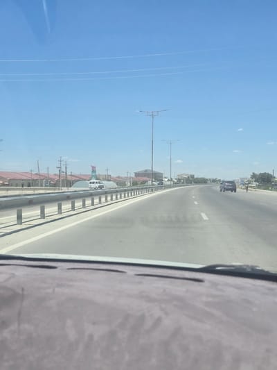 A wide-angle, medium-distance shot captures a bright, sunny day on a multi-lane road in Yukari-Gazbiran, Uzbekistan. The road is paved and appears to be an expressway or highway, with clear lane markings. A metal guardrail runs along the left side of the road, separating it from the adjacent area. On the left, a few low-rise buildings with red roofs are visible, along with power lines and utility poles. A white van is parked near one of these buildings, and a single person is walking in the distance. On the right side of the road, a dark-colored SUV is driving away from the camera, and a person on a bicycle is also moving along the roadside. In the distance, more buildings and trees can be seen, and a signpost is visible on the right. The sky is a clear, vibrant blue with faint contrails from an aircraft. The foreground shows the dashboard of a vehicle, indicating the photo was taken from inside a car. The overall scene depicts a typical day on a road in a semi-urban or suburban environment, with vehicles and pedestrians navigating the infrastructure under a clear sky.