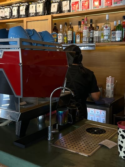 Here is a detailed description of the image:
The image shows a cafe or bar setting, potentially located in Mazatlán, Mexico, given the Spanish product labels. A person in a black uniform is working at an espresso machine. The machine is red and has a stack of blue coffee cups attached to its side. Behind the person and the espresso machine, there is a shelf with various bottles of alcohol, including brands like Bacardi and Jim Beam. There are also packages of coffee beans labeled "estelar" on the shelf. Cartons of "Santa Clara" milk are visible above the espresso machine. A metal grate with a cup rinser is located on the right. The general atmosphere suggests a commercial establishment preparing beverages.