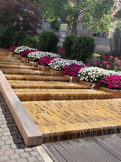 The image depicts a tiered water fountain in Levico Terme, Italy. The fountain features multiple levels of warm-toned, textured stone steps, with water cascading down each level. Several small fountains are active on the stone surfaces, creating sprays of water. Along the top edge of the fountain, rows of vibrant pink and white impatiens flowers are planted in raised beds, contrasting with the stone and water. Green bushes are situated behind the flowers. In the background, a large tree with lush green leaves provides shade, and a red triangular road sign with black arrows is visible. To the right, a portion of a dark statue of a seated figure with braided hair can be seen, and further back, a set of wide stone stairs leads upwards, bordered by trees and buildings. The foreground includes a textured cobblestone path on the left and a wooden deck on the right. The lighting suggests it is a sunny day.