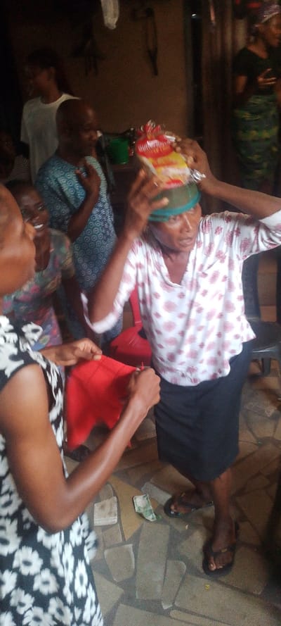 This image captures an indoor gathering or celebration in what appears to be a home or community space in Ohafia, Nigeria. The scene is lively, with several individuals present, their expressions suggesting joy and engagement.
In the center, an older woman wears a white patterned blouse, a dark skirt, and a light green headscarf. Her hands are raised to her head as someone places a clear plastic-wrapped package, likely a snack or bread item, on top of her head. Partial text visible on the packaging includes "BUTTER" and "FLAVOUR." Her expression is focused, perhaps reacting to the item being placed on her. She wears black flip-flops.
To the left in the foreground, a woman in a black and white floral patterned dress holds a folded red piece of fabric, seemingly in the process of handing it over or receiving it. Behind her, another woman is smiling broadly, and a man in a blue patterned shirt is also visible, smiling with his hands slightly raised. Further back on the right, another woman in a dark top and patterned wrapper skirt watches the activity, also appearing to smile. The background shows parts of a dark wall, a green bucket, and a red plastic chair.
On the patterned tile floor, a crumpled banknote, likely Nigerian Naira, is visible, along with another piece of paper. The overall atmosphere suggests a moment of celebration, gift-giving, or a traditional practice, characterized by interaction and positive emotions among the participants. The lighting indicates an indoor setting, possibly in the evening or a dimly lit room.