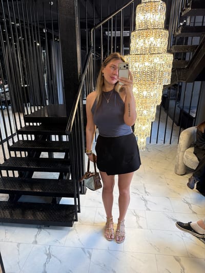 A woman is taking a selfie in a modern, upscale interior, possibly a boutique or salon, with a large, multi-tiered crystal chandelier and a black spiral staircase. She is wearing a grey halter top and a black wrap skirt, with studded gold sandals. She is holding a brown and light-colored handbag. Her left arm has a tattoo. The floor is tiled with a white marble pattern. A portion of a gray armchair and the feet of another person wearing black Crocs and white socks are visible to the right. The background features black metal railings and possibly changing room curtains. The location is not specified as being in Укромновское сельское поселение, Ukraine.