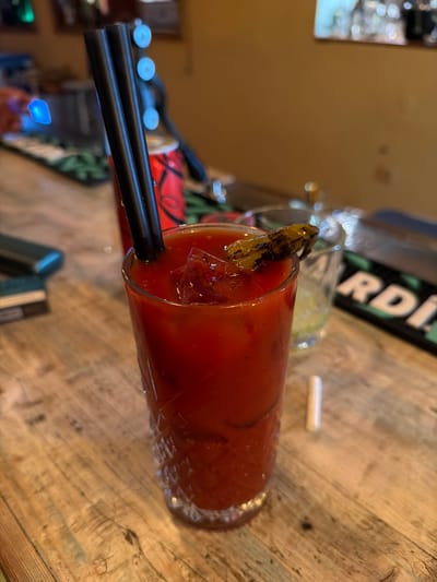 A tall glass of red cocktail with ice and two black straws is placed on a wooden bar. A small green garnish sits on the rim of the glass. In the background, a red can and other drinks are visible. The lighting is dim, suggesting an indoor setting. The image appears to be taken in Varvara, Bulgaria, at a bar.