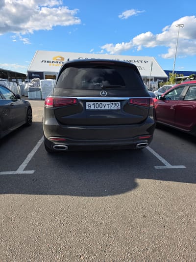 This media file shows the rear of a dark-colored Mercedes-Benz SUV parked in an outdoor lot. The vehicle has a license plate that reads "B 100 УТ 790 RUS". The background includes a building with signage that says "ЛЕМ", "КЕНТР", and "Вход", suggesting it might be a store or business center. Another red car is partially visible to the left. The scene appears to be a sunny day, as indicated by the bright sky with scattered clouds and the distinct shadow cast by the Mercedes. Parking lot markings are visible on the asphalt. The overall setting is a commercial area, likely a shopping center or a business park, under a clear sky. The text on the building and the car's license plate suggest the location is in Russia. No people are visible in the frame.