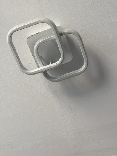 This is a close-up, overhead shot of two interlocking, square-shaped metal light fixtures mounted on a textured white ceiling. The fixtures are painted white and cast subtle shadows on the ceiling, suggesting a light source from above and slightly to the left. The texture of the ceiling is uneven, with visible grain and some faint marks or imperfections. The overall impression is minimalist and modern. The image does not contain any people, text, or explicit location cues beyond what might be inferred from the style of the fixtures or the typical interior of a building. The time of day and weather are not discernible.