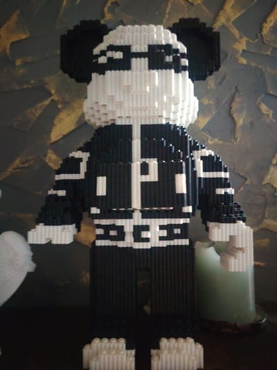 A tall, black and white pixelated figure resembling a panda wearing sunglasses and a jacket is positioned centrally against a textured, abstract background. The figure is constructed from numerous small, cylindrical blocks, creating a distinct pixelated aesthetic. To the left of the figure, a partial view of a white, wing-like object is visible. To the right, a large, pale green candle in a dish is present. The overall scene appears to be an indoor display, possibly in a home or a gallery setting, in the city of Sukharevo, Russia. The lighting is somewhat dim, with the main light source appearing to be from above and slightly to the right.