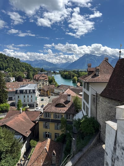 A high-angle shot captures the city of Thun, Switzerland, on a bright, sunny day. The foreground is dominated by the terracotta-tiled roofs of historic buildings, characteristic of the region. A cobblestone pathway winds its way down, bordered by a stone wall and lush green foliage. Several people are visible walking down this path, dressed in casual attire. In the mid-ground, a turquoise river, likely the Aare River, flows through the city, flanked by more buildings with traditional architecture. Further in the background, rolling green hills covered in trees lead to majestic snow-capped mountains under a sky filled with dramatic white clouds against a vibrant blue backdrop. The overall scene depicts a charming European city nestled between a river and the Alps.