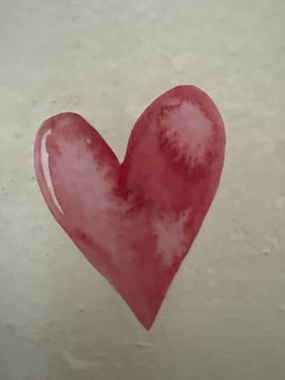 The image shows a watercolor painting of a pink heart on a textured, light-colored background. The heart is depicted with soft edges and variations in color intensity, suggesting a watercolor technique. There are no people or discernible objects that would indicate a specific location in Ushtobe, Kazakhstan. The image is a close-up of the artwork itself.