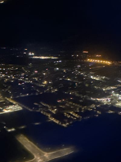 The aerial photograph captures a nighttime view of a city, likely from an airplane. The scene is dominated by darkness, with illuminated urban features standing out against the black sky. Numerous lights, appearing as blurry dots and streaks of varying colors, indicate buildings, streets, and possibly vehicles. The lights are concentrated in certain areas, suggesting the presence of a city or town.
In the foreground and extending into the middle ground, there appears to be a body of water, possibly a river or a bay, reflecting some of the artificial lights. Along the edge of this water, illuminated structures and what could be port facilities or a harbor are visible.
Further inland, the landscape is dotted with clusters of lights, hinting at residential areas, commercial districts, and industrial zones. Some areas show a higher density of lights, suggesting more developed sections of the city, while others appear sparser. There are distinct lines of lights that could represent roads or highways.
The overall impression is one of a vast, sprawling urban area at night, seen from a significant altitude. The lack of sharp detail due to the nighttime conditions and possible motion blur from the camera's perspective adds to the abstract quality of the image.
While the provided image itself does not offer explicit geographical markers to definitively confirm the location as Surcin Urban Municipality, Serbia, it depicts a general urban landscape at night, consistent with what might be observed over such an area from an aerial viewpoint.