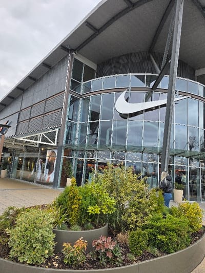 This image captures the exterior of a Nike retail store located within an outdoor shopping complex in Zweibrücken, Germany, specifically identifying with the Zweibrücken Fashion Outlet. The environment is depicted under an overcast sky.

In the foreground, a large, curved, multi-tiered concrete planter is filled with a variety of shrubs and small plants, exhibiting green, yellow-green, and reddish-brown foliage. The ground beneath is paved with light-colored pavers.

The dominant structure is a modern commercial building featuring a curved glass facade on its lower and middle sections. A large, white Nike swoosh logo is prominently displayed centrally on the glass. The upper portion of the building showcases a dark, patterned wall texture and is topped by a wide, grey, angled roof supported by exposed dark grey steel beams and structural elements. Through the glass, interior lighting, merchandise displays, and a large graphic image of a person are discernible.

One individual is visible on the right side of the image, standing near the building's facade and facing towards its entrance. This person has blonde, shoulder-length hair and is wearing a black puffer jacket and blue jeans. Additional blurred figures are observable inside the store through the glass windows. Two light-colored, cylindrical potted plants are positioned near the building's facade on the right.
