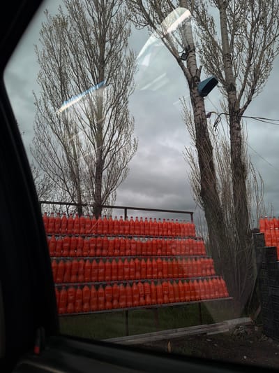 The image shows a view through a car window in the city of Gagarin, Armenia. The background consists of an overcast sky and two bare-branched trees. A rack filled with rows of orange plastic bottles is situated between the trees. Additionally, a small, blue object is attached to one of the tree branches. In the upper right corner are more black crates.