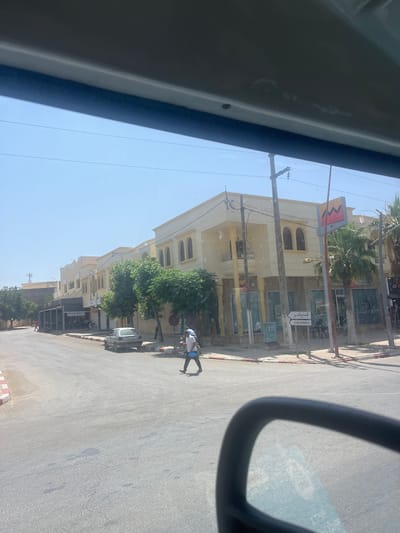 The image, taken from inside a vehicle, shows a street scene in Boufakrane, Morocco. A pale yellow building with arched windows lines the street. A street sign with Arabic and French text points to "EL HAJJ KADDOUR". A silver car is parked on the side of the road. A man is crossing the street in the foreground. Another man is walking a bicycle on the right side of the frame. The sky is clear and blue.