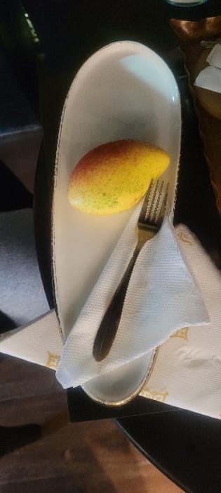 A mango sits on a white, elongated plate trimmed with gold, next to a folded white napkin and a fork. The mango is yellow with tinges of red and slight green speckling. The fork rests partially on the mango and the napkin. The setup is on a dark surface, partially reflecting light, and a light-colored garment is visible underneath the plate. A decorative element is also seen in the background.