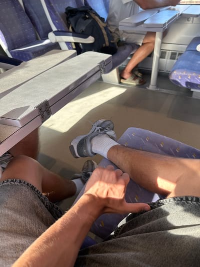 The image shows two people's legs on a train. The person in the foreground is wearing grey denim shorts and a grey t-shirt, with their right arm visible, adorned with fine hairs. Their left hand is giving a thumbs-up gesture. They are sitting on a purple seat. The person in the background is wearing grey sneakers and white socks, with one leg extended. They appear to be sitting on a similar purple seat. The train carriage has blue and purple patterned seats and metal accents on the tables. Sunlight streams in, casting shadows and highlighting the textures of the denim and hair. The overall scene suggests a casual journey on a sunny day in Rivesaltes, France. There are no visible signs of distress or negative emotions; the atmosphere seems relaxed and positive, indicated by the thumbs-up. The setting is clearly an indoor train compartment.