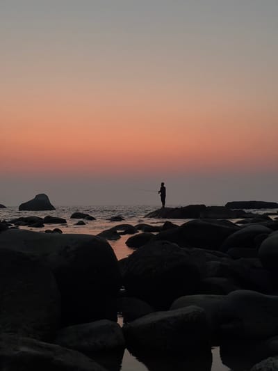 A silhouetted figure stands fishing near a rocky shoreline at dusk. The sky transitions from light gray to warm orange, reflecting faintly on the water. Dark, rounded rocks line the right side of the frame, creating a stark contrast with the lighter horizon and fisherman. The composition is vertical, emphasizing the transition in colors.
