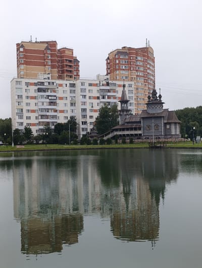 This is a wide, overcast, eye-level outdoor shot of a tranquil pond and its reflection, with a large residential building complex and a wooden church visible in the background. The pond is in the foreground, taking up roughly the bottom half of the frame, and its surface is calm, creating a clear reflection of the scene above. The reflection shows the multi-story residential buildings, the church, trees, and the cloudy sky.
In the middle ground, on the far side of the pond, stands a large, modern residential building complex. It appears to be composed of at least two connected towers. The tower on the left is shorter and has a facade primarily of light beige brick with darker red brick accents on its upper levels. The tower on the right is taller and more curved, with a similar brick pattern of alternating beige and red sections, interspersed with numerous windows. In front of these towers, and slightly to the right, is a traditional-looking wooden church. The church is grey, made of logs, and features multiple onion domes, a bell tower, and a covered walkway. A staircase leads up to the church's entrance.
Behind the church and extending to the right are dense green trees, forming a natural backdrop. To the left of the residential complex, there are also trees and some signs of a street or pathway with lampposts. The sky is uniformly grey, indicating an overcast day. The overall impression is a juxtaposition of modern urban architecture with historical religious structures, set against a natural element of water. The location is identified as Kiyevsky, Russia.