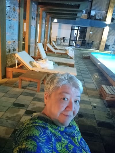 The image depicts an outdoor scene at what appears to be a resort or a recreational area. In the foreground, a person is taking a selfie, their face and upper body visible. They have short, graying hair and are wearing a blue shirt with a green, tropical leaf pattern. The background shows a walkway paved with large rectangular tiles. To the left, there are several empty lounge chairs with white cushions arranged under a covered walkway supported by wooden pillars. Decorative panels with intricate patterns are mounted on the walls of this walkway. Further to the right, a bright blue swimming pool is visible. Wooden crates are placed near the pool and along the walkway. The environment appears to be lit by artificial lights, suggesting it might be evening or night. The overall atmosphere suggests a relaxing or holiday setting. The location in the city of Sorbulak, Kazakhstan, is stated, implying this scene is situated within that area.