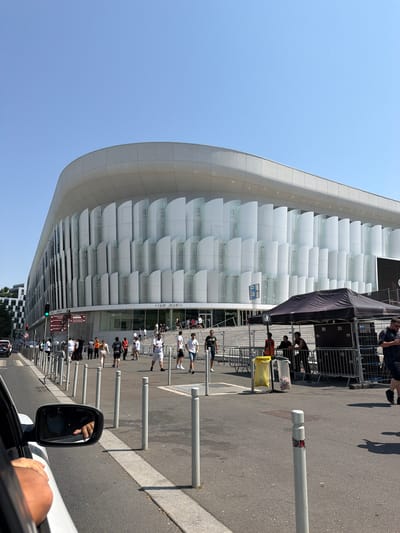 The image shows a wide, curved, modern building with a white, textured facade featuring vertical, translucent panels. In front of the building, a street is visible with a sidewalk and a road. Several people are walking along the sidewalk, some near a black tent set up with metal barriers. A yellow trash can and a clear plastic bag filled with trash are also present. Traffic lights and street signs are visible on the left side of the image. The sky is clear and blue, indicating a sunny day. The scene is likely in the city of Nanterre, France, as suggested by the presence of street signs with French text and the architectural style, possibly near a stadium or large venue. The perspective is from inside a vehicle, with a hand visible on the window frame and a side mirror reflecting the interior.