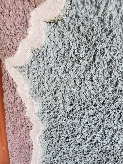 The image displays a close-up, abstract view of a textured wall. The wall has three distinct sections: on the left, a rough, reddish-purple surface; in the middle, a wavy line of light pink material; and on the right, a predominantly gray, speckled, and rough texture. The lighting suggests an outdoor environment, possibly daylight, casting subtle shadows that highlight the wall's uneven surface. There are no people, animals, or clear objects depicted in the frame, making it difficult to ascertain a specific location within Konstantinovka, Moldova, or any discernible action or interaction. The focus is solely on the material and texture of the wall.