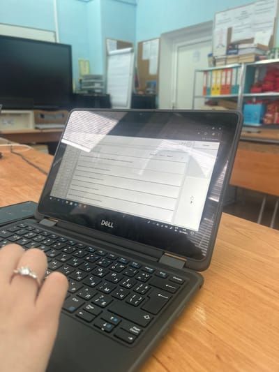 The image depicts an interior scene, likely a classroom or administrative office, within the city of Dolni Tsibar, Bulgaria. In the foreground, a left hand with a silver ring on the fourth finger is positioned over the black QWERTY keyboard of a Dell laptop, which also features Cyrillic characters. The laptop screen displays a document or web page with multiple text fields and questions in Bulgarian, indicating an active task, possibly a test or questionnaire. Horizontal reflections of light are visible across the laptop screen. The laptop rests on a light-colored wooden desk.

In the blurred background to the left, a large, dark flat-screen monitor sits on a lower desk, with a white printer or scanner unit positioned behind it. The wall in this area is light blue. To the right in the background, a shelving unit holds various items, including multiple colored binders (yellow, orange, red, green), stacks of papers or books, and several red and blue packaged items. Above this shelving unit, a bulletin board with pinned papers is visible. Further back, a white door frame and another bulletin board or whiteboard are partially obscured. The overall environment suggests an educational or institutional setting. The laptop's system tray shows the date as 20.12.2020.