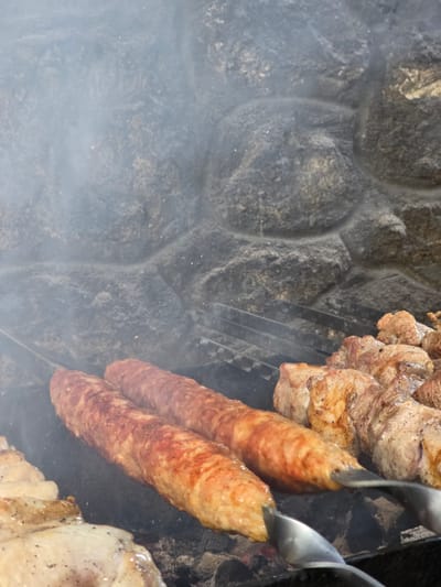 In Pidhorodne, Ukraine, a close-up shot captures two long, sausage-like kebabs cooking on a grill. The kebabs, seasoned and browned, are skewered and positioned prominently in the foreground. Behind them, pieces of cubed meat, also on skewers, are being grilled, their surfaces showing signs of charring and seasoning. Wisps of smoke rise from the grill, indicating active cooking. The background features a rough, stone-like surface, possibly part of an outdoor grill or a rustic wall, adding a natural and earthy element to the scene. The lighting is dim, suggesting it might be evening or an overcast day, with the focus on the food and the cooking process.
