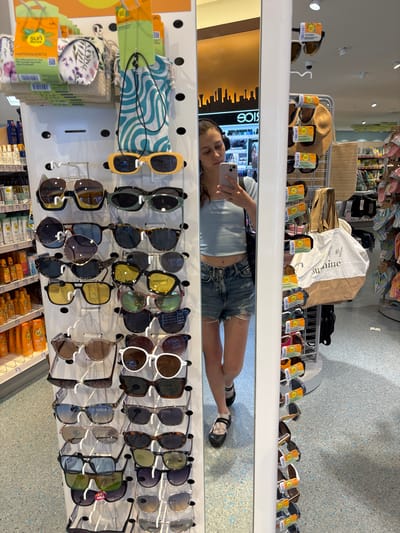 This media file captures a reflection in a store mirror, where a young woman is taking a selfie. She is wearing a light blue tank top and distressed denim shorts, and she has brown hair. The store appears to be a retail environment selling sunglasses, hats, and beach accessories. Various pairs of sunglasses are displayed on shelves, along with protective cases and a straw hat. The store has a bright, clean aesthetic with white shelving units and a speckled grey and blue wall.
The reflection also shows a display of brightly colored flip-flops and other items on a higher shelf. In the foreground of the reflection, we see shelves stocked with what appear to be sunscreen bottles and small bags. The overall impression is of a sunny day, likely in the summer, given the products on display and the woman's attire. The woman's expression is neutral, as she focuses on taking her photograph. The visible text includes brand names and product descriptions, such as "SUN", "BRILLENKETTE", and "HARTSCHALENETUR", which are German for "sunglasses chain" and "hard shell case", respectively. The location is identified as Erlenbach am Main, Germany.