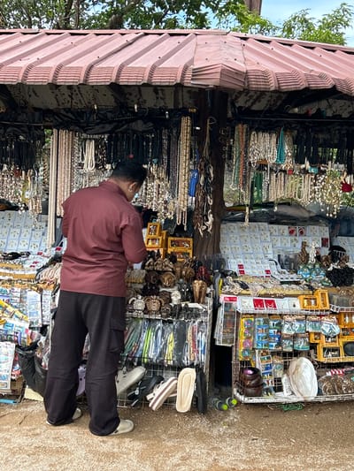 A man wearing a maroon long-sleeved shirt and black pants stands in front of a market stall displaying numerous necklaces and jewelry. The stall has a reddish-brown corrugated metal roof supported by wooden posts. Necklaces of various sizes, colors, and materials hang from hooks. Various small items, including what appear to be carved wooden animals, are also displayed. In the foreground, there are several pairs of sandals and flip-flops on the ground. The ground is covered with gravel. The scene appears to be outdoors with foliage visible in the background. The image is taken from a slightly low angle.
